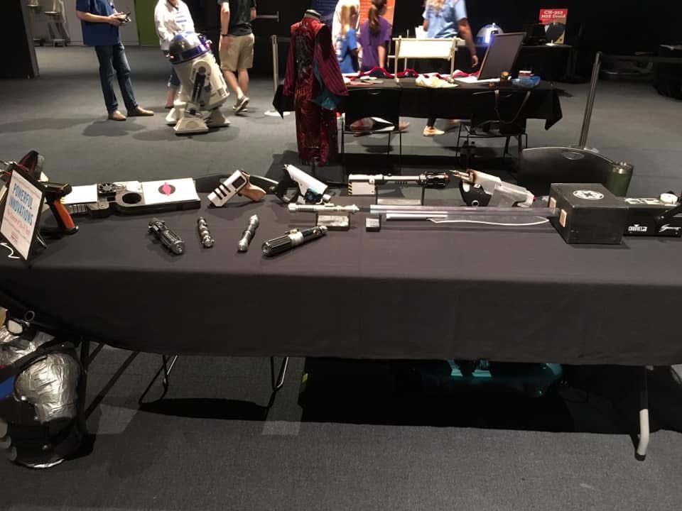 My table at the Science Center of Iowa maker fair: lightsabers, laser builds and props laid out on a black tablecloth, with an R2-D2 style droid and visitors behind